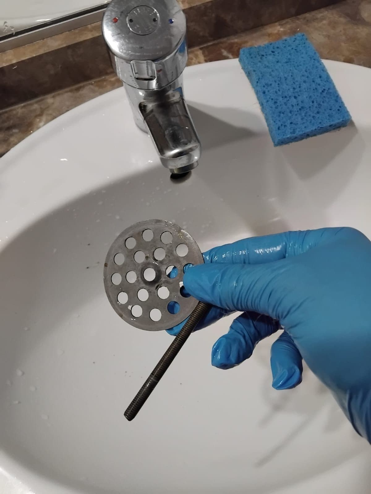 Grille de bonde démontée tenue au-dessus d'un lavabo, gant bleu et éponge